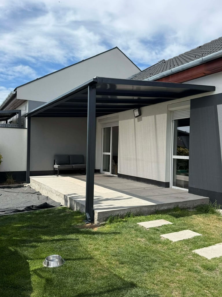 Modern gray house extension with black pergola covering a paved patio area and manicured lawn